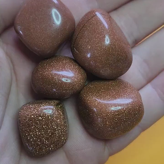 Hand holding gold sandstone tumbles against a yellow background, sparkling glittery man-made goldstone crystals for confidence, motivation, and energy healing, close-up product video
