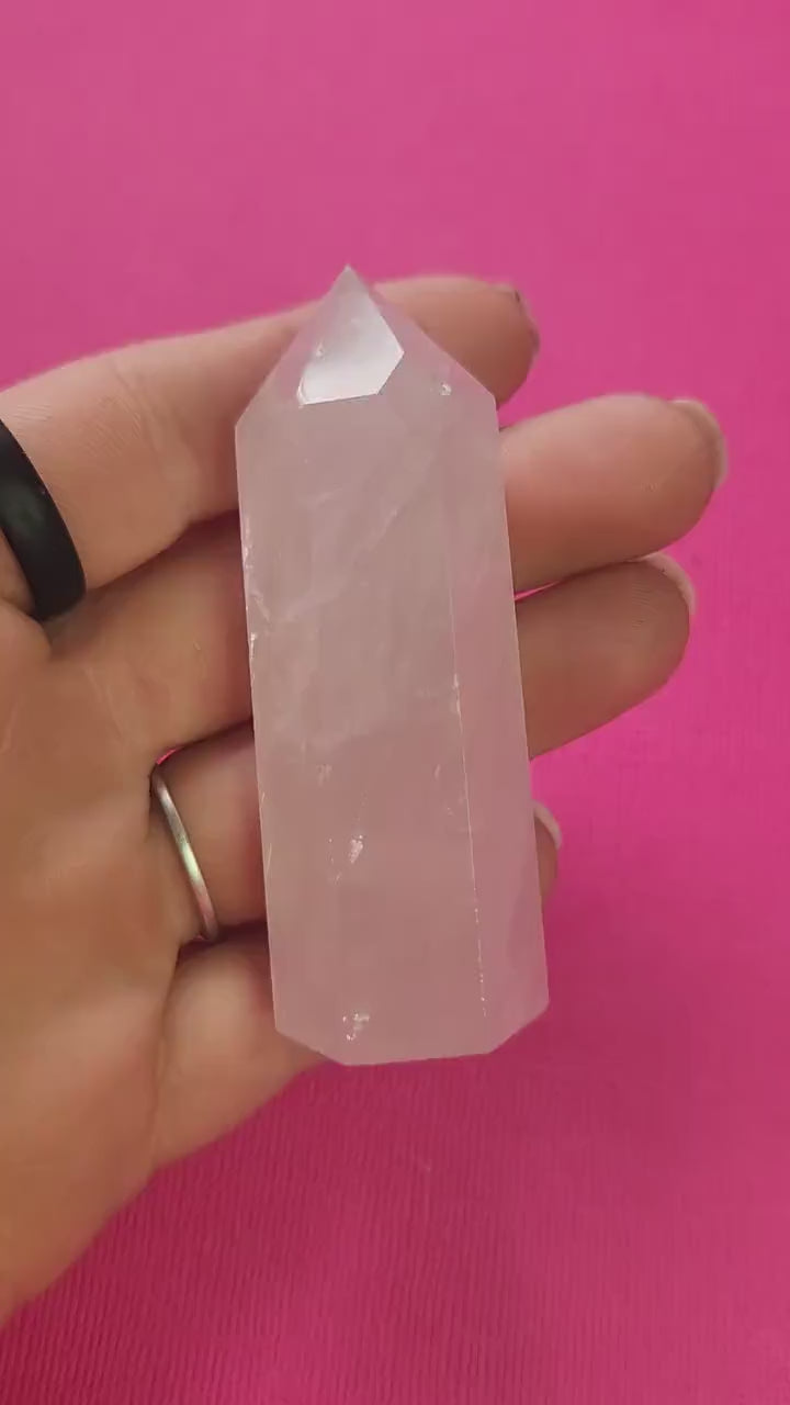 Video of a hand holding and turning a polished rose quartz crystal tower against a bright pink background, showing its soft pink hue and glossy finish. The gentle lighting highlights the stone’s natural beauty and soothing, heart-healing energy — perfect for self-love and emotional balance.