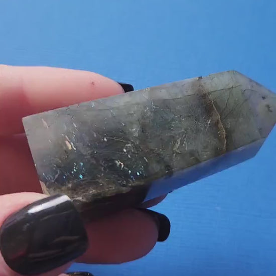Close-up of a hand holding a polished Labradorite tower showing vivid blue flashes against a blue backdrop. Natural Labradorite crystal tower ideal for meditation, aura protection, Chakra healing, energy work, Reiki, spiritual practice, home décor, and crystal collections. Perfect for crystal enthusiasts, intuitive guidance, and thoughtful gifts.