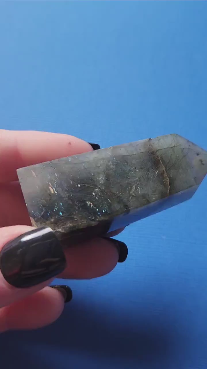 Close-up of a hand holding a polished Labradorite tower showing vivid blue flashes against a blue backdrop. Natural Labradorite crystal tower ideal for meditation, aura protection, Chakra healing, energy work, Reiki, spiritual practice, home décor, and crystal collections. Perfect for crystal enthusiasts, intuitive guidance, and thoughtful gifts.