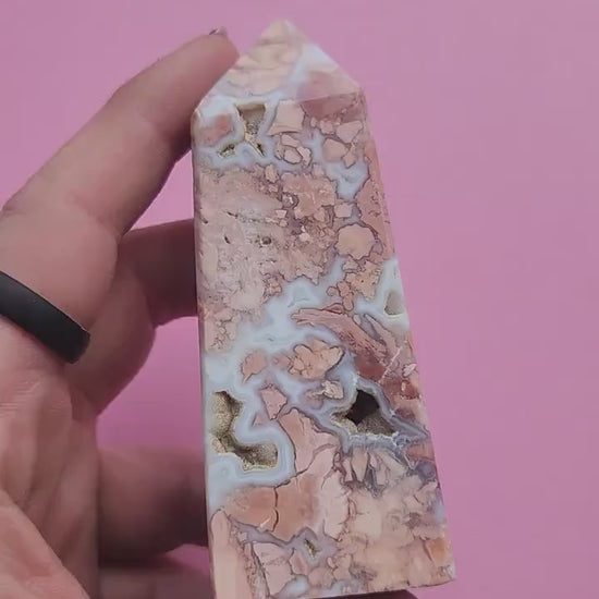 Hand holding a pink cotton candy agate crystal tower against a soft pink background showing pastel banding and polished agate point for crystal healing decor metaphysical crystals and gift