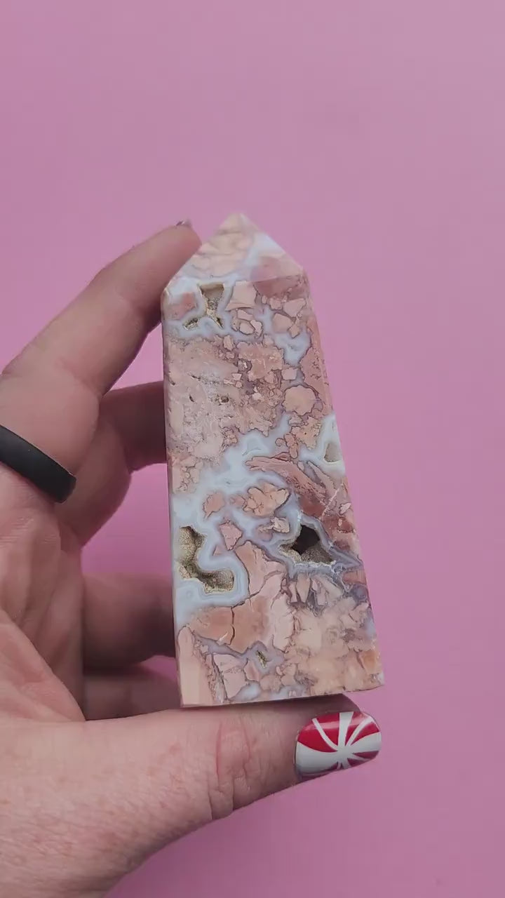 Hand holding a pink cotton candy agate crystal tower against a soft pink background showing pastel banding and polished agate point for crystal healing decor metaphysical crystals and gift
