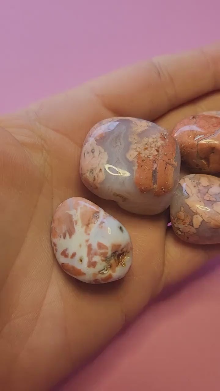 Hand holding Cotton Candy Agate tumbled stones, also known as Pink or Sakura Agate, against a pink background, showcasing polished pink gemstones for emotional healing, calming energy, self-love, and crystal collection display.