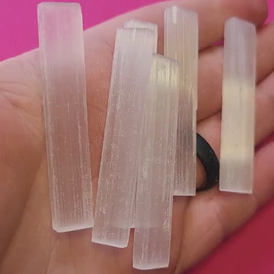Video of hand holding a raw Selenite Satin Spar stick against a bright pink background. The white, fibrous crystal glows softly in the light. Perfect for cleansing energy, spiritual healing, and crystal collections