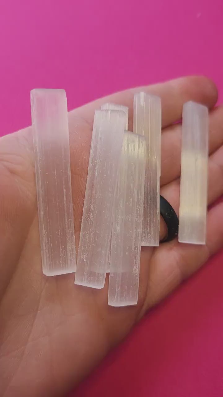 Video of hand holding a raw Selenite Satin Spar stick against a bright pink background. The white, fibrous crystal glows softly in the light. Perfect for cleansing energy, spiritual healing, and crystal collections
