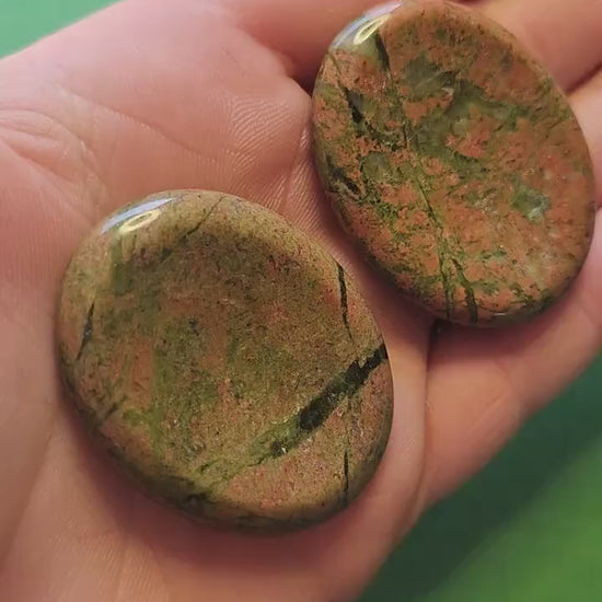 Video of two Unakite worry stones held in hand against a green background, showing pink and green polished stones — nurturing and balancing crystal for emotional healing, harmony, and grounding.