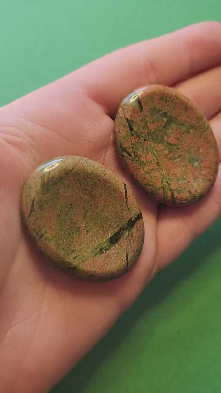 Video of two Unakite worry stones held in hand against a green background, showing pink and green polished stones — nurturing and balancing crystal for emotional healing, harmony, and grounding.