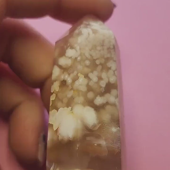Video of a hand holding a Flower Agate tower against a light pink background, showcasing its soft pink and white crystal tones with natural floral-like inclusions, polished finish, and pointed tower shape — ideal for healing, meditation, and crystal display.