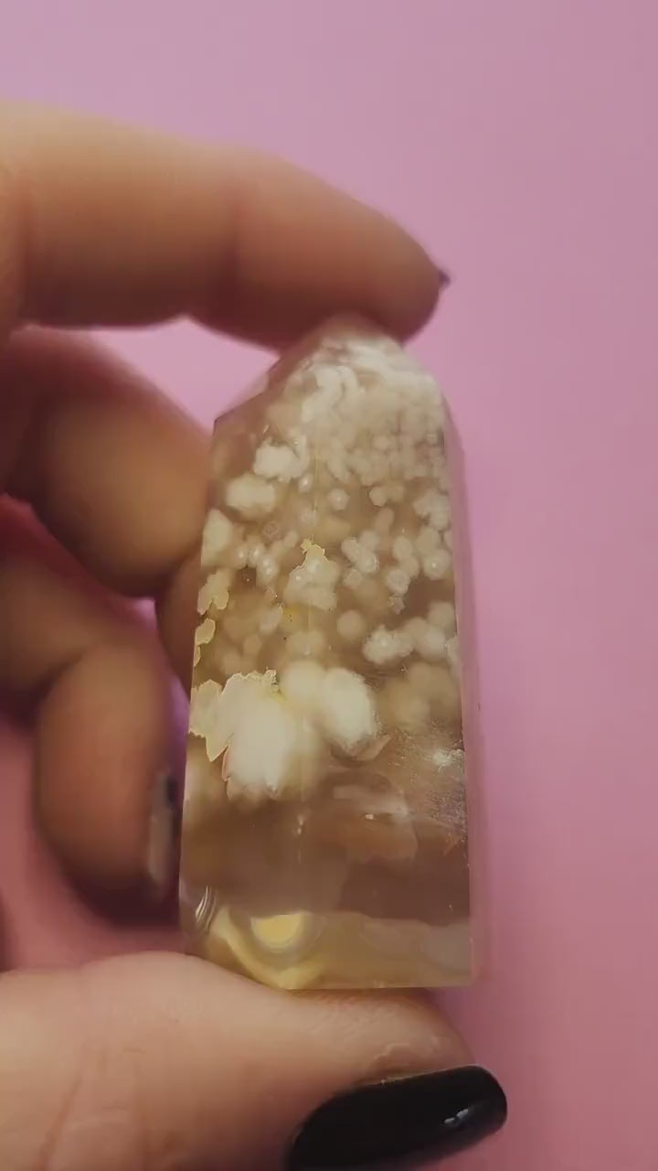 Video of a hand holding a Flower Agate tower against a light pink background, showcasing its soft pink and white crystal tones with natural floral-like inclusions, polished finish, and pointed tower shape — ideal for healing, meditation, and crystal display.