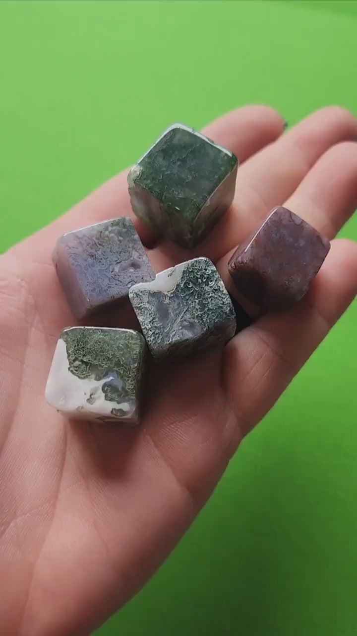 Close-up video of a hand holding a polished Moss Agate cube against a green background. Each natural cube (approx. 1.7–2cm) features translucent to milky white chalcedony with deep green moss-like inclusions. Perfect for grounding, meditation, crystal grids, heart chakra work, spiritual décor, and as a meaningful gift for crystal collectors.