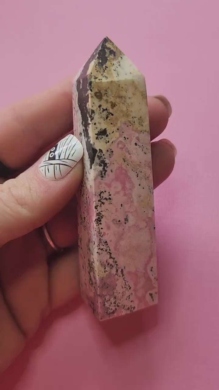 Video of Peruvian Rhodonite tower held in hand against pink background, showing polished pink and black banding, high-quality rhodonite crystal point for self-love, emotional healing and heart chakra support.