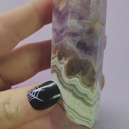Video shows a hand holding an Amethyst and Crazy Lace Agate crystal tower up close against a soft lavender background. The camera focuses on the tower’s unique mix of purple amethyst and banded agate patterns, highlighting its natural shimmer and detail. The lighting accentuates the crystal’s texture and color variations, making it perfect for showcasing gemstone quality and beauty.