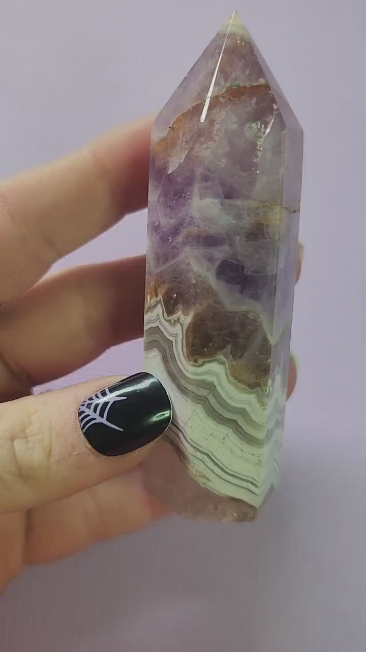 Video shows a hand holding an Amethyst and Crazy Lace Agate crystal tower up close against a soft lavender background. The camera focuses on the tower’s unique mix of purple amethyst and banded agate patterns, highlighting its natural shimmer and detail. The lighting accentuates the crystal’s texture and color variations, making it perfect for showcasing gemstone quality and beauty.