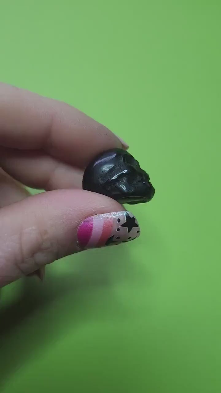 Close-up video of a hand holding a mini Black Obsidian crystal skull with a vibrant green background, showcasing polished surface and detailed carving. Perfect for protection, shadow work, Halloween decor, and carved crystal skull collectors.