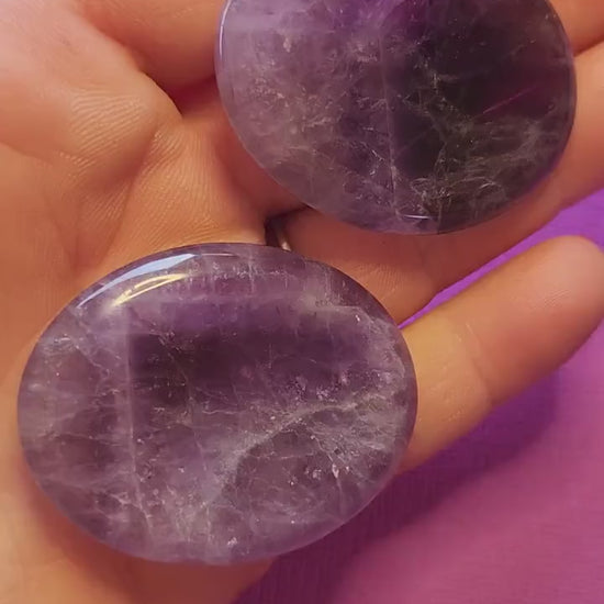 Polished Amethyst thumb stone held in hand, close-up view against a purple background. Smooth purple crystal for stress relief, meditation, spiritual awareness, Third Eye balance, and inner peace.