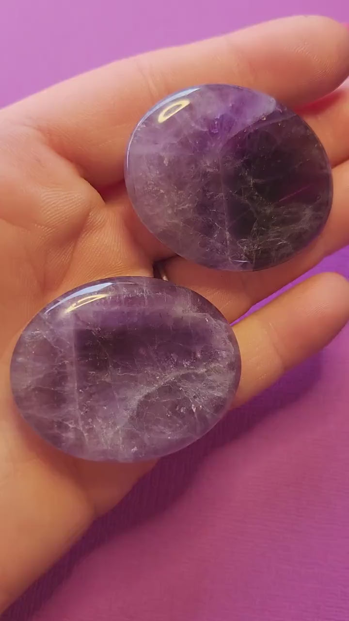 Polished Amethyst thumb stone held in hand, close-up view against a purple background. Smooth purple crystal for stress relief, meditation, spiritual awareness, Third Eye balance, and inner peace.