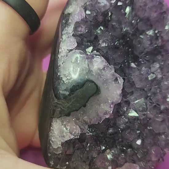 Video shows a hand holding an Amethyst cut base crystal up close against a rich purple background. The camera highlights the sparkling purple points, natural druzy texture, and polished edges of the geode. Lighting enhances the depth of color and clarity, showcasing the crystal’s beauty, quality, and vibrant healing energy.
