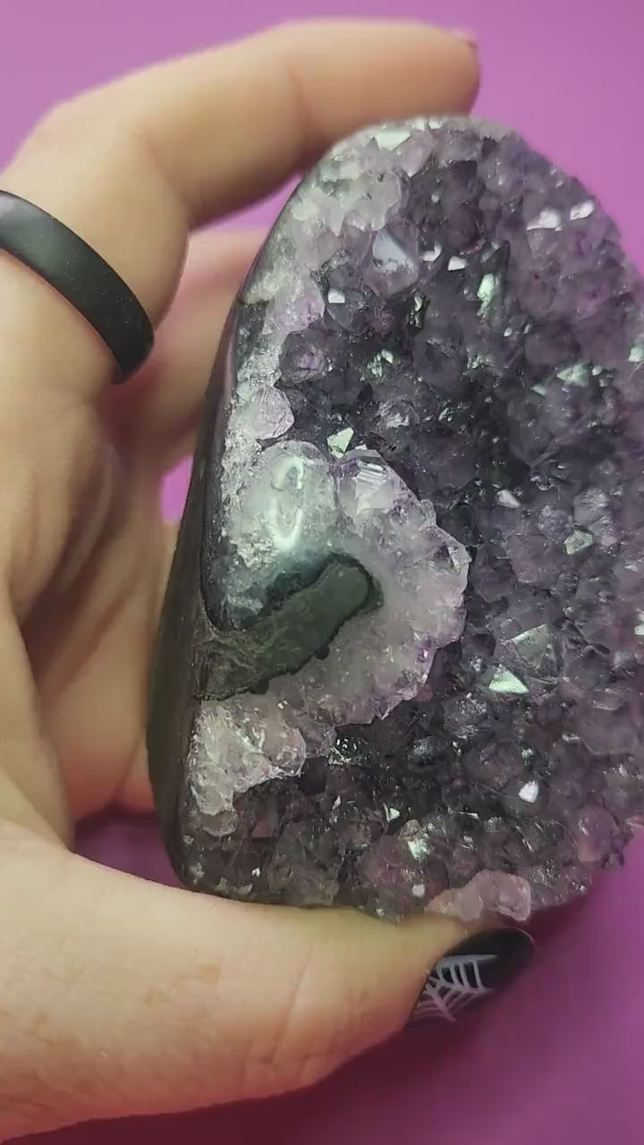 Video shows a hand holding an Amethyst cut base crystal up close against a rich purple background. The camera highlights the sparkling purple points, natural druzy texture, and polished edges of the geode. Lighting enhances the depth of color and clarity, showcasing the crystal’s beauty, quality, and vibrant healing energy.