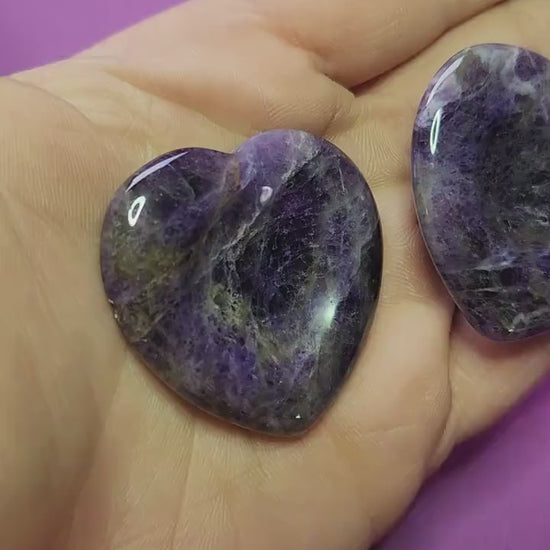 Showing two Amethyst heart-shaped worry stones in my hand against a soft purple background, close-up clip highlighting their polished purple crystal surface and soothing stress-relief energy, no props.