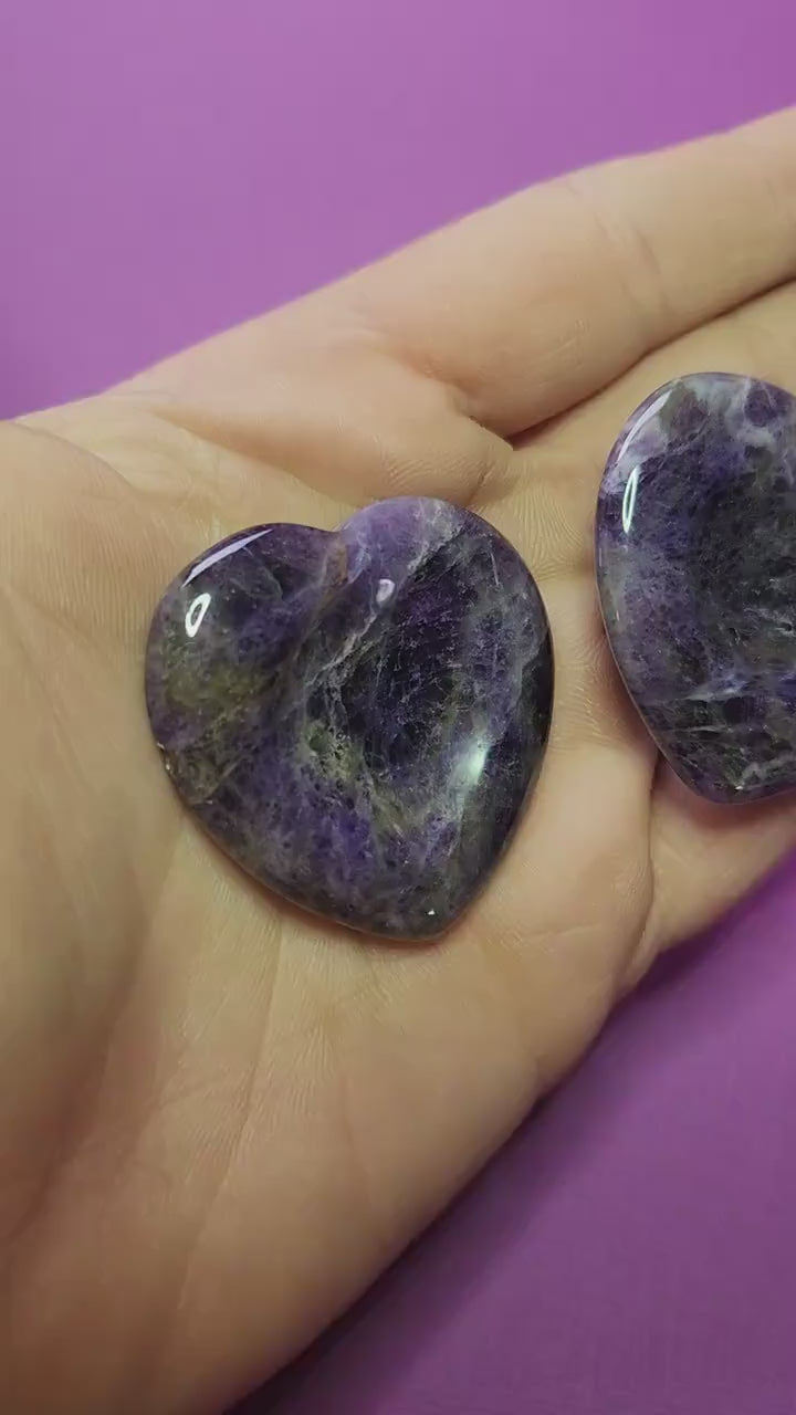 Showing two Amethyst heart-shaped worry stones in my hand against a soft purple background, close-up clip highlighting their polished purple crystal surface and soothing stress-relief energy, no props.