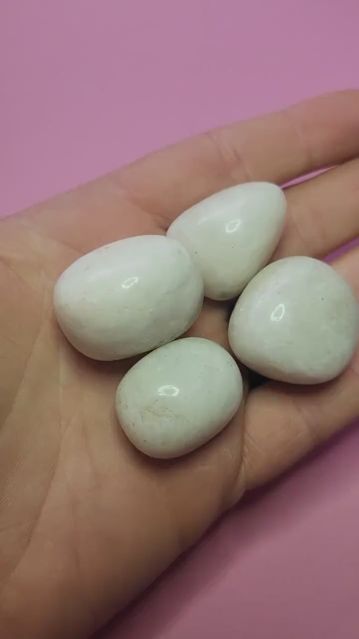 Close-up video of polished White Moonstone crystal tumbles held in hand against a pink background, showcasing natural moonstone shimmer and iridescence, ideal for intuition enhancement, emotional balance, feminine energy, calming energy, meditation, chakra healing, manifestation, spiritual crystal collections, and metaphysical décor
