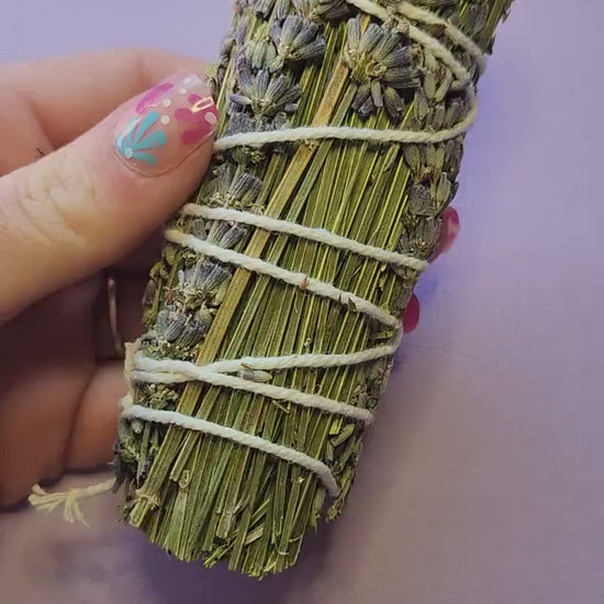 Hand holding a natural Lavender Smudge Stick against a lavender background, featuring dried lavender bundles for smudging, cleansing negative energy, aromatherapy, meditation, relaxation, spiritual rituals, and energy clearing.