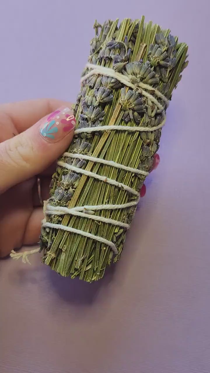 Hand holding a natural Lavender Smudge Stick against a lavender background, featuring dried lavender bundles for smudging, cleansing negative energy, aromatherapy, meditation, relaxation, spiritual rituals, and energy clearing.