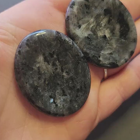 Two polished Larvikite worry stones shown up close in hand, featuring dark grey color with silver-blue flashes. Grey backdrop, perfect for grounding, protection, meditation, and crystal healing.