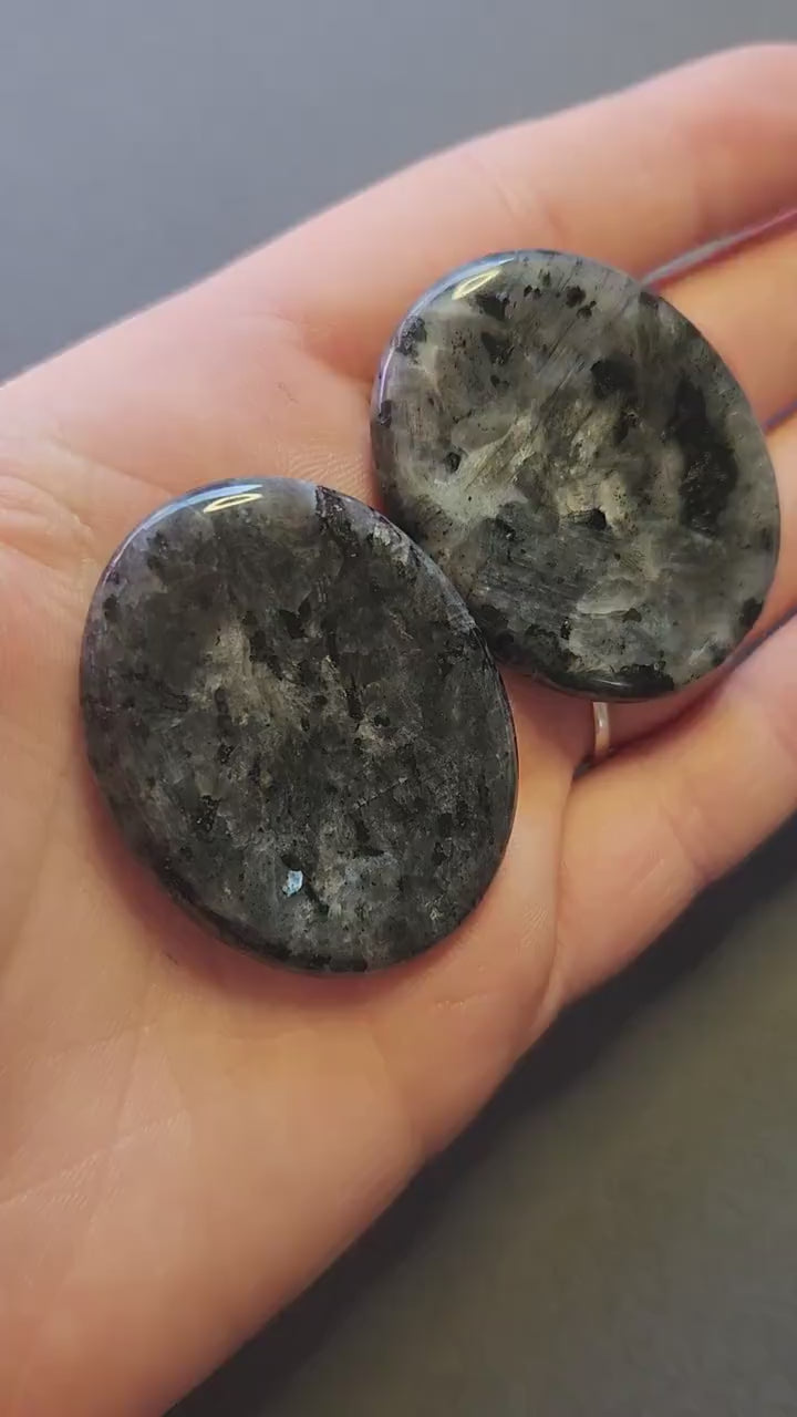 Two polished Larvikite worry stones shown up close in hand, featuring dark grey color with silver-blue flashes. Grey backdrop, perfect for grounding, protection, meditation, and crystal healing.