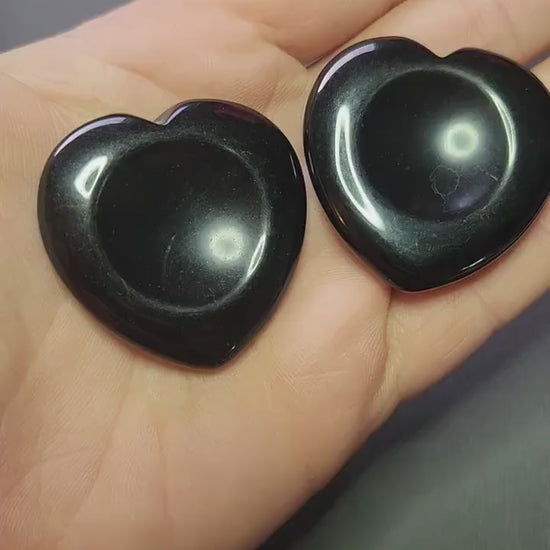 Showing two black obsidian heart-shaped worry stones in my hands against a dark grey background, polished black obsidian hearts for protection, grounding and negative energy cleansing, crystal worry stones close-up for shop showcase.