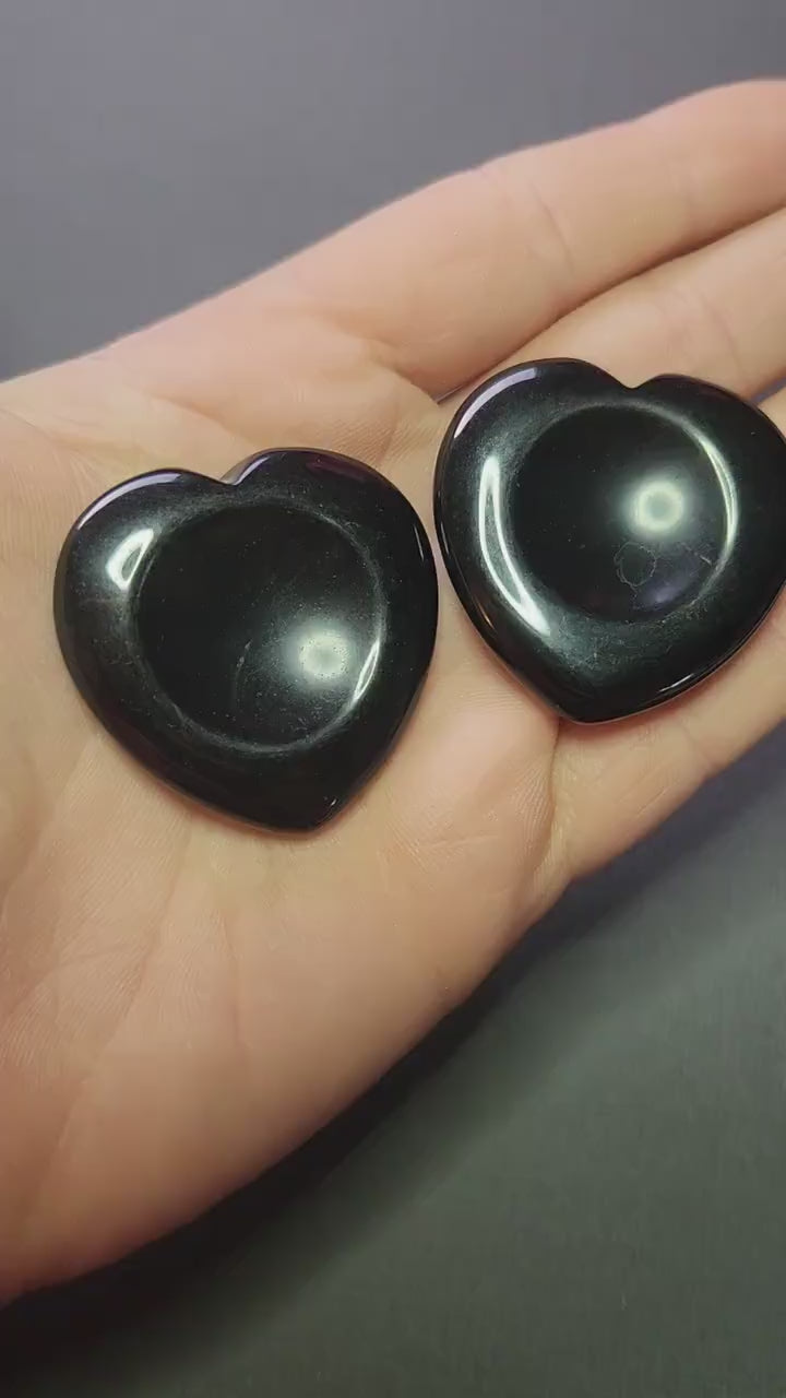 Showing two black obsidian heart-shaped worry stones in my hands against a dark grey background, polished black obsidian hearts for protection, grounding and negative energy cleansing, crystal worry stones close-up for shop showcase.