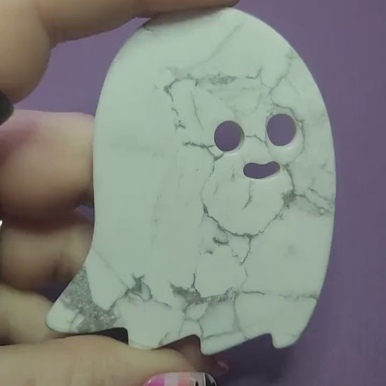 Close-up of a hand holding a carved white Howlite ghost slice with natural grey veining against a dark purple backdrop. Flat, palm-sized ghost crystal perfect for Halloween altars, witchy décor, crystal grids, meditation, calming energy, spiritual practice, and spooky-season gifts.