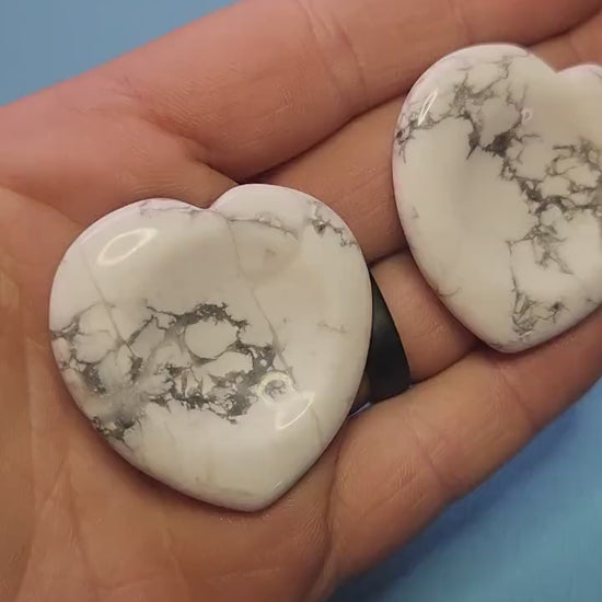 Two howlite heart-shaped worry stones held in hand against a light blue background, white calming crystals for anxiety relief, howlite heart worry stones for stress soothing, crystal shop showcase close-up.