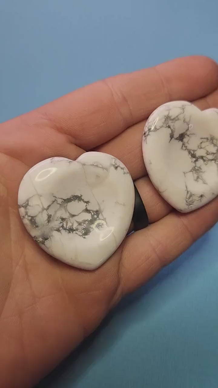 Two howlite heart-shaped worry stones held in hand against a light blue background, white calming crystals for anxiety relief, howlite heart worry stones for stress soothing, crystal shop showcase close-up.
