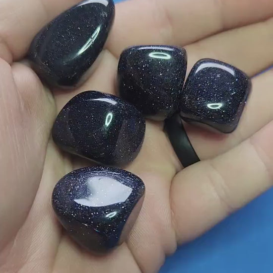 Close-up video of polished Blue Sandstone crystal tumbles held in hand against a blue background, showcasing natural deep blue shimmer and sparkling inclusions, ideal for grounding energy, calming energy, meditation, chakra balancing, manifestation, spiritual crystal collections, and metaphysical décor