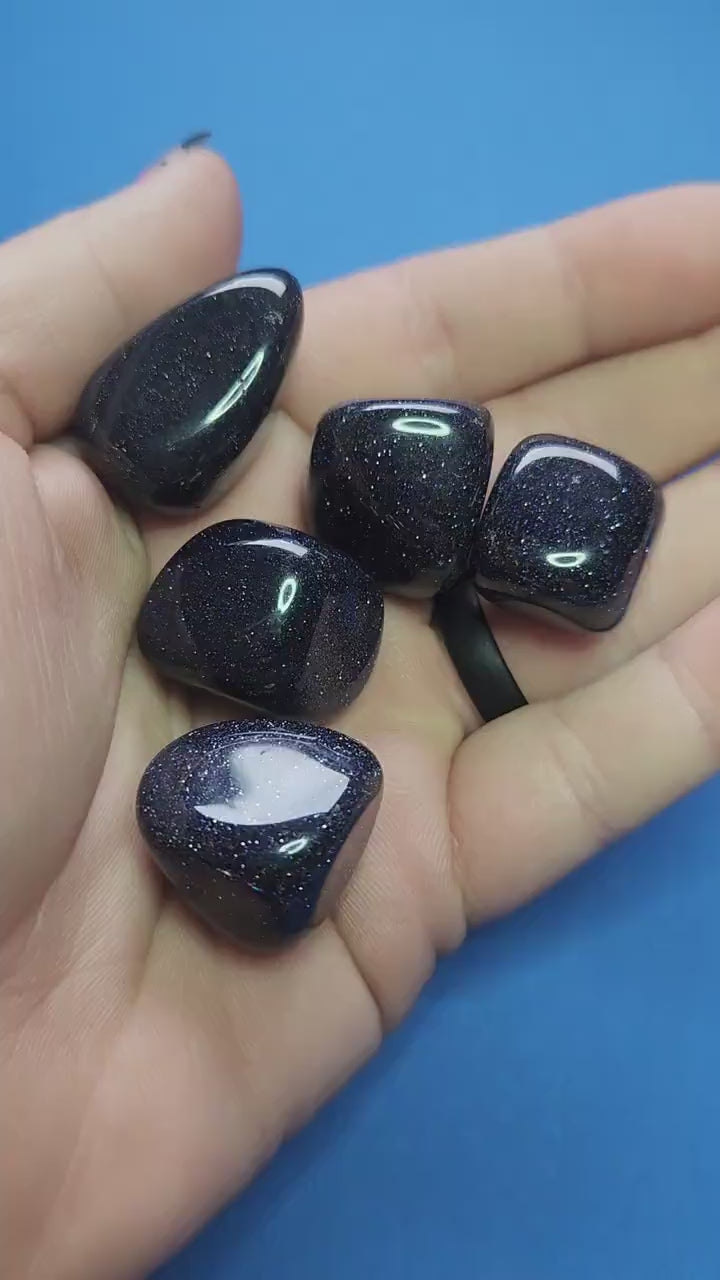 Close-up video of polished Blue Sandstone crystal tumbles held in hand against a blue background, showcasing natural deep blue shimmer and sparkling inclusions, ideal for grounding energy, calming energy, meditation, chakra balancing, manifestation, spiritual crystal collections, and metaphysical décor