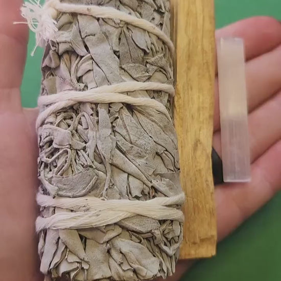 Close-up video of a hand holding a cleansing bundle featuring a white sage stick, palo santo stick, and selenite stick against a green backdrop, showcasing the natural textures and sizes of each item — ideal for spiritual cleansing and energy healing enthusiasts.