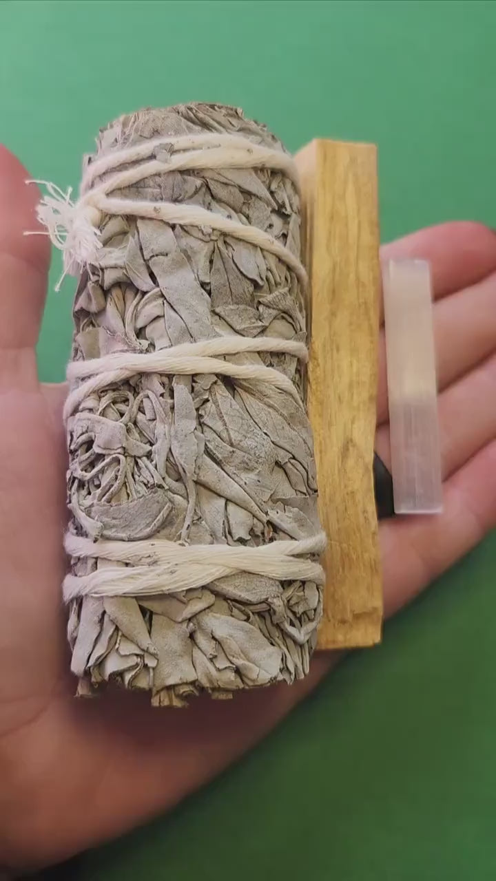 Close-up video of a hand holding a cleansing bundle featuring a white sage stick, palo santo stick, and selenite stick against a green backdrop, showcasing the natural textures and sizes of each item — ideal for spiritual cleansing and energy healing enthusiasts.