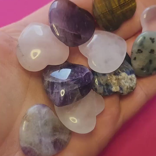 Hand holding assorted small crystal hearts on a soft pink background, showcasing polished stones like rose quartz, amethyst, and aventurine.