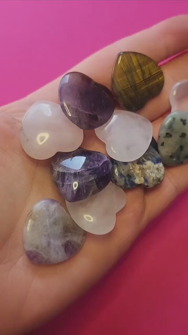 Hand holding assorted small crystal hearts on a soft pink background, showcasing polished stones like rose quartz, amethyst, and aventurine.