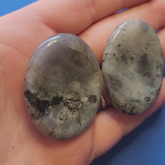 Polished Labradorite worry stone held in hand against a blue background, showing flashes of blue, green, and gold. Smooth thumb stone for meditation, mindfulness, aura protection, stress relief, and crystal healing. Ideal for pocket talismans, spiritual gifts, and grounding practices.