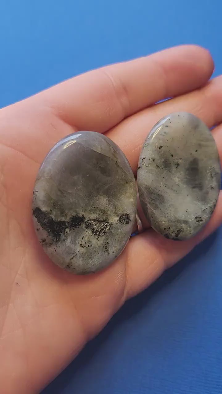 Polished Labradorite worry stone held in hand against a blue background, showing flashes of blue, green, and gold. Smooth thumb stone for meditation, mindfulness, aura protection, stress relief, and crystal healing. Ideal for pocket talismans, spiritual gifts, and grounding practices.