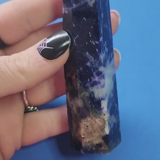 Close-up video of a polished Sodalite crystal tower held in hand against a blue background, showcasing deep blue color with white veining, ideal for intuition enhancement, mental clarity, meditation, chakra balancing, manifestation, spiritual crystal collections, metaphysical décor, and high-vibration crystal healing practices
