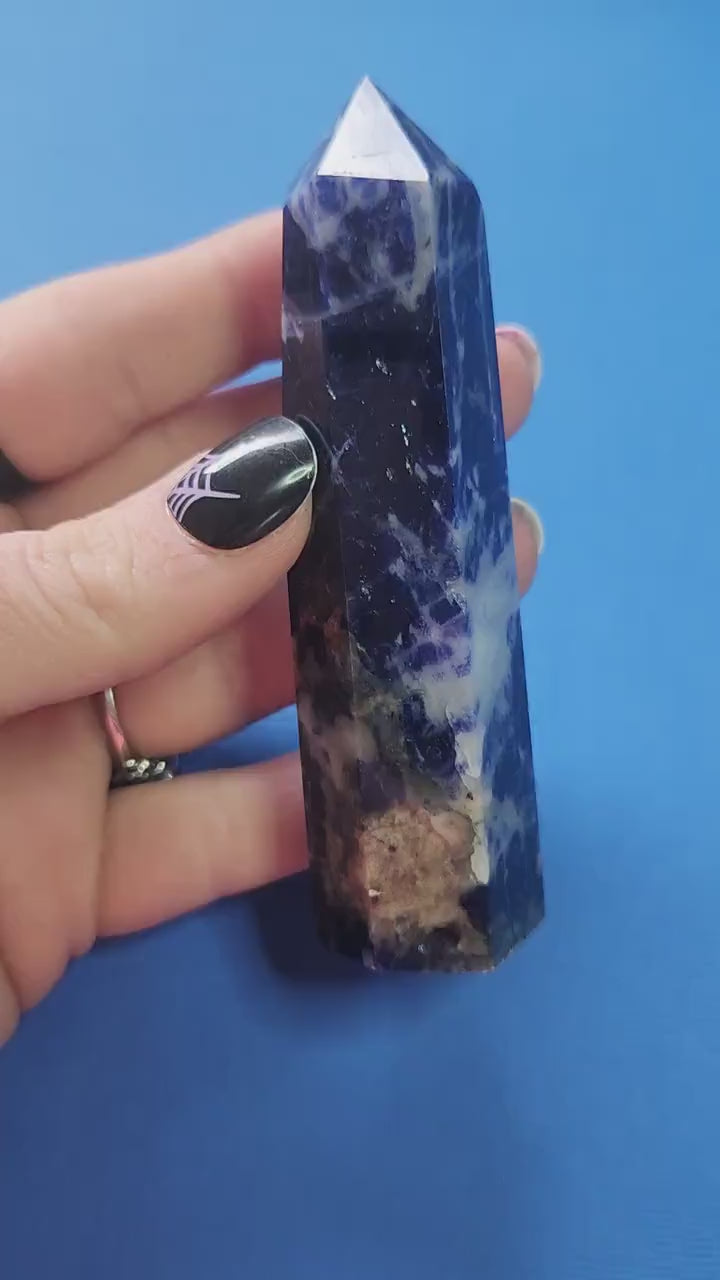 Close-up video of a polished Sodalite crystal tower held in hand against a blue background, showcasing deep blue color with white veining, ideal for intuition enhancement, mental clarity, meditation, chakra balancing, manifestation, spiritual crystal collections, metaphysical décor, and high-vibration crystal healing practices