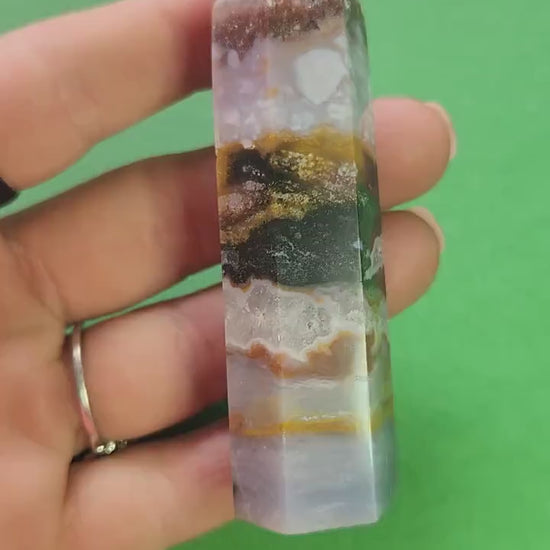 Video of Ocean Jasper tower held in hand against green background, showing polished orbicular patterns in green, cream and brown, natural gemstone point for grounding, emotional healing, meditation, and heart chakra support