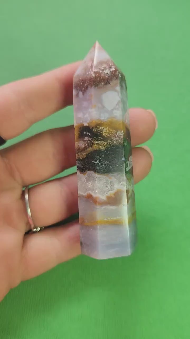 Video of Ocean Jasper tower held in hand against green background, showing polished orbicular patterns in green, cream and brown, natural gemstone point for grounding, emotional healing, meditation, and heart chakra support