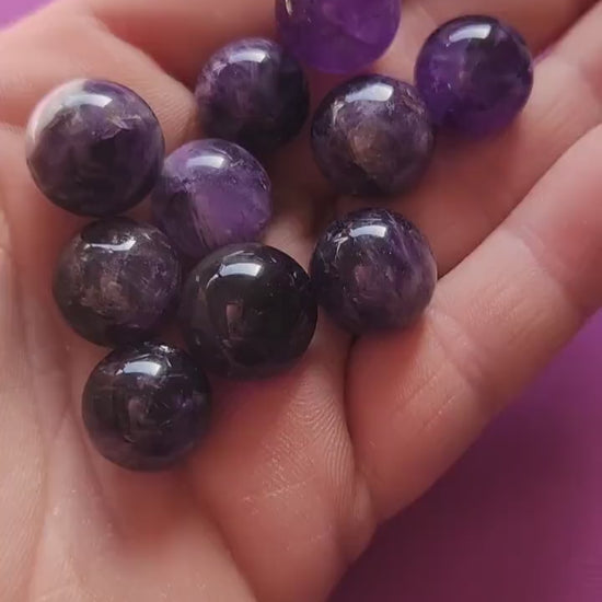 Hands holding and showcasing polished Amethyst mini spheres against a dark purple backdrop, highlighting their smooth texture and deep violet hues.