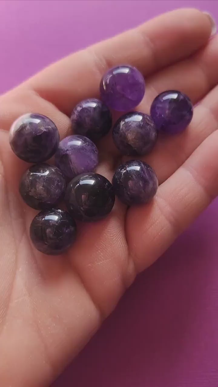Hands holding and showcasing polished Amethyst mini spheres against a dark purple backdrop, highlighting their smooth texture and deep violet hues.