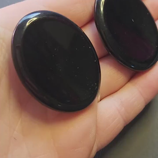 Polished Black Obsidian thumb stone held in hand, close-up view against a dark grey background. Pocket protection crystal for grounding, emotional balance, shielding energy, and spiritual practice.