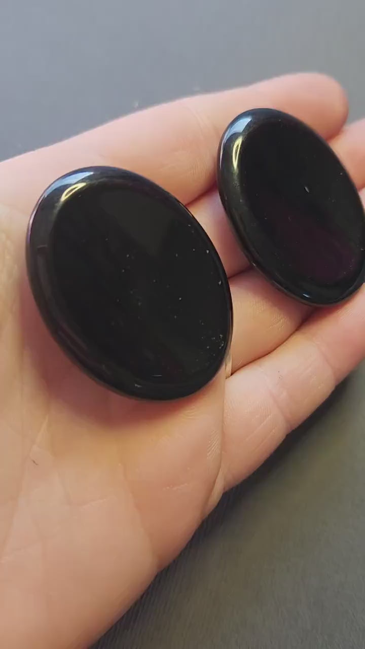 Polished Black Obsidian thumb stone held in hand, close-up view against a dark grey background. Pocket protection crystal for grounding, emotional balance, shielding energy, and spiritual practice.